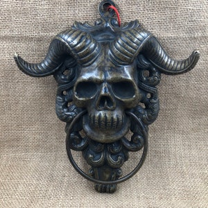 以下が含まれることがあります： ブロンズ色のスカルドアノッカー。湾曲した角とリング状のハンドルが付いています。スカルには詳細な眼窩、鼻腔、顎があります。ノッカーは、装飾的な裏板に装飾的なスクロールワークと吊り下げ用のループが付いています。