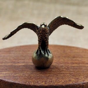Peut inclure: Une figurine d'aigle en bronze aux ailes déployées, perchée sur une petite sphère. Les ailes de l'aigle sont détaillées et la sculpture a un aspect vieilli. La figurine est exposée sur une surface en bois.