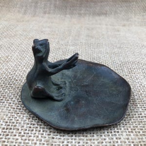 Könnte beinhalten: Eine bronzene Froschfigur sitzt auf einer schalenförmigen Seerosenblatt. Der Frosch hat die Arme erhoben, und die Schale hat eine strukturierte Oberfläche. Die Figur ist ein dekoratives Objekt.