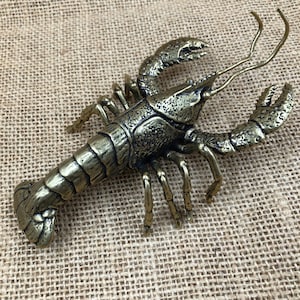 Puede incluir: Figura de langosta de metal color bronce con detalles texturizados en el cuerpo, pinzas y patas. Las antenas de la langosta son largas y curvadas. La figura se exhibe sobre una superficie texturizada de color beige.