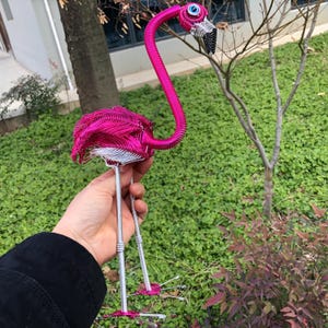 May include: A handcrafted, wire flamingo sculpture in vibrant pink. The bird features a long, curved neck, detailed wings, and slender legs. The flamingo's eye is a blue bead, and the beak is black. The sculpture is held against a backdrop of green foliage.