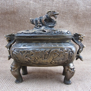 May include: Bronze incense burner with detailed dragon motifs. The lid is topped with a dragon sculpture, and the body features dragon designs. It has three ornate legs. A decorative item for home interiors, showcasing detailed craftsmanship.