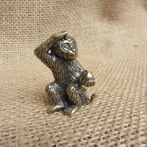Peut inclure: Petite figurine de singe en bronze, assis et se grattant la tête d'une main, tenant un petit objet dans l'autre. Le singe a une texture de fourrure détaillée et une expression souriante.