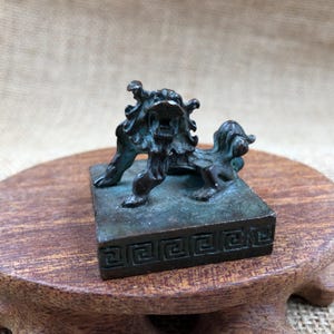 May include: A bronze-colored, small statue of a mythical lion-like creature. The statue is mounted on a square base with a Greek key design. The creature has a fierce expression with its mouth open, and its fur is detailed.