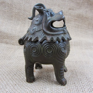 May include: A bronze-colored, decorative incense burner in the shape of a mythical creature. The detailed sculpture features a lion-like head with an open mouth, intricate spiral patterns, and a textured mane. The burner stands on four short legs.