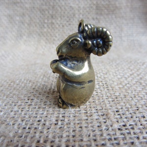 May include: A small, detailed brass figurine of a ram, standing upright with its front hooves clasped together. The ram has large, curled horns and a loop at the top for hanging. The figurine has a vintage, antique appearance.