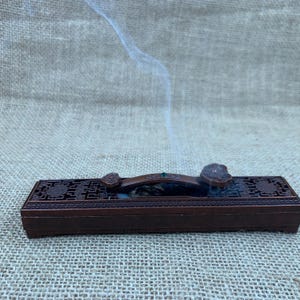 May include: A rectangular, dark brown metal incense burner box featuring an intricate, openwork lattice pattern on the lid. A curved, decorative handle with a small green accent sits atop the lid, with a thin trail of white smoke rising upwards.