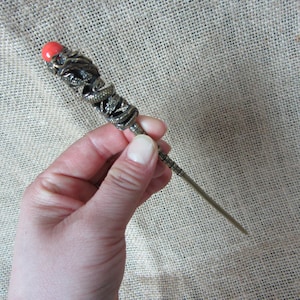 Peut inclure: Une épingle à cheveux en bronze antique avec un motif de dragon. Le dessus présente un motif de dragon détaillé et une perle rouge. L'épingle est tenue dans une main sur un fond neutre.