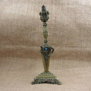 May include: An ornate bronze-colored dagger with intricate detailing. The dagger features a triangular blade, a decorative handle, and a turquoise-colored stone. The base is square with floral patterns. The overall design suggests a ceremonial or decorative item.
