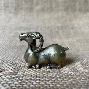 Peut inclure: Une petite figurine métallique de couleur laiton représentant un bélier en position de repos. La sculpture présente des cornes incurvées détaillées, un museau défini et un corps lisse et arrondi, capturant l'animal assis, pattes repliées.