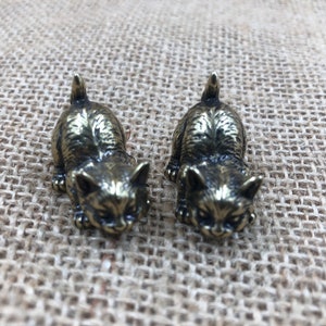 May include: Two small, bronze-colored cat figurines. The cats are in a crouching position, with their tails raised. The figurines have detailed features, including ears, eyes, and paws. The background is a textured, tan fabric.