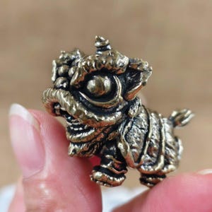 May include: A small, detailed brass figurine of a mythical lion-like creature. The figurine has intricate carvings, a textured surface, and a weathered, antique finish. The creature is in a crouching position, with its head raised.