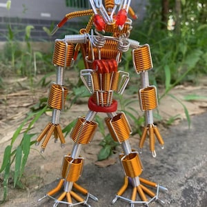 Peut inclure: Une sculpture de robot faite à la main, fabriquée à partir de fil de couleur cuivre et de fil argenté. Le robot présente une tête et une poitrine rouges, avec du fil enroulé formant le corps, les bras et les jambes. Le robot est posé sur une surface en béton.