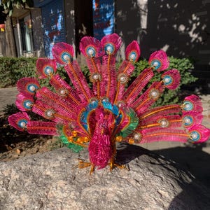 May include: A vibrant, handcrafted peacock sculpture made of pink wire. The peacock's tail feathers are fanned out, showcasing intricate details with blue and green accents, and faux pearls. The sculpture is perched on a gray rock, with a sunny outdoor background.