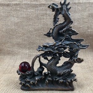 Könnte beinhalten: Bronzene Drachenskulptur mit einer roten Glaskugel. Der Drache ist aufwendig detailliert, mit Schuppen und fließenden Merkmalen. Die Skulptur ist auf einem Sockel in Form von Meereswellen montiert.