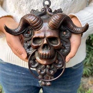 May include: A bronze-toned metal door knocker featuring a detailed human skull with large, curved ram horns. The design includes ornate, swirling scrollwork surrounding the skull, with a circular metal ring suspended from the jaw.
