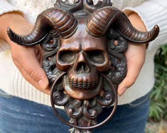 May include: A bronze-toned metal door knocker featuring a detailed human skull with large, curved ram horns. The design includes ornate, swirling scrollwork surrounding the skull, with a circular metal ring suspended from the jaw.