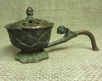 May include: An antique bronze incense burner with a lotus flower design. The burner has a lid with small holes and a handle. The base is a detailed floral pattern. The color is a mix of bronze and green.