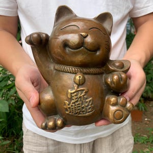 Könnte beinhalten: Bronzestatue einer Maneki Neko Katze mit erhobener Pfote und einer Goldmünze. Die Katze hat einen lächelnden Ausdruck und einen dekorativen Kragen mit einer Glocke und chinesischen Schriftzeichen. Die Statue wird vor einem grünen Hintergrund gehalten.