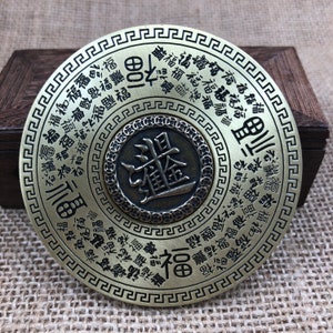 Könnte beinhalten: Eine runde, antik-messingfarbene Metallscheibe mit chinesischen Schriftzeichen. Die Scheibe hat konzentrische Kreise mit Text und einem zentralen Design. Der Text enthält das Zeichen für "Glück" und andere Symbole. Die Scheibe wird auf einer strukturierten Oberfläche präsentiert.