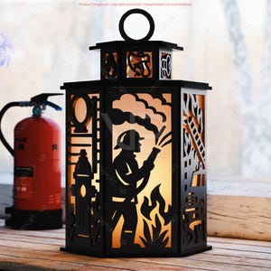 May include: Black decorative lantern with a firefighter silhouette design. The lantern features cut-out details of a firefighter, fire, ladder, and fire hydrant. The lantern has a handle and is placed on a wooden surface.