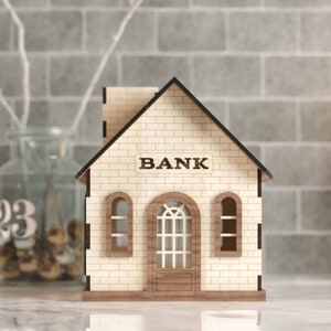 以下が含まれることがあります： 「BANK」と書かれた看板のある木製の銀行の建物の模型。模型には窓が2つとドアが1つある。建物は白い表面の上に置かれている。