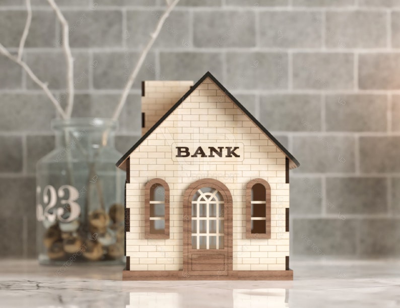 以下が含まれることがあります： 「BANK」と書かれた看板のある木製の銀行の建物の模型。模型には窓が2つとドアが1つある。建物は白い表面の上に置かれている。