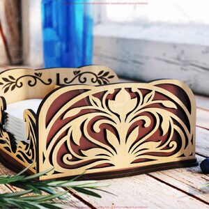 Pu&ograve; includere: Porta tovaglioli in legno con intricati ritagli floreali. Il supporto presenta un esterno marrone chiaro e un interno marrone scuro, e contiene una pila di tovaglioli bianchi. Il design include motivi decorativi di foglie e volute.