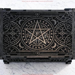 May include: A black wooden box with intricate silver-colored engravings. The design features a central pentagram, crescent moons, stars, and floral patterns. The box has a rectangular shape with detailed corner joints.