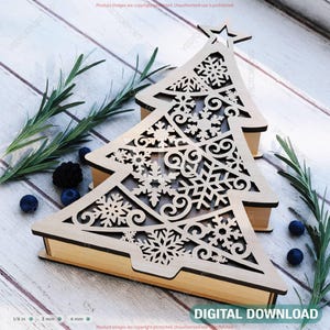 Puede incluir: Caja de madera con forma de árbol de Navidad con intrincados recortes de copos de nieve. La caja es de color marrón claro con una estrella en la parte superior. La imagen también incluye ramitas de romero, arándanos y el texto "DIGITAL DOWNLOAD".