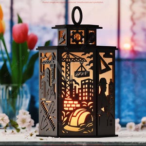 May include: Black laser-cut lantern with construction-themed cutouts, including a crane, buildings, and a hard hat. The lantern has a handle and is illuminated from within, creating a warm glow. The background features flowers and a window.