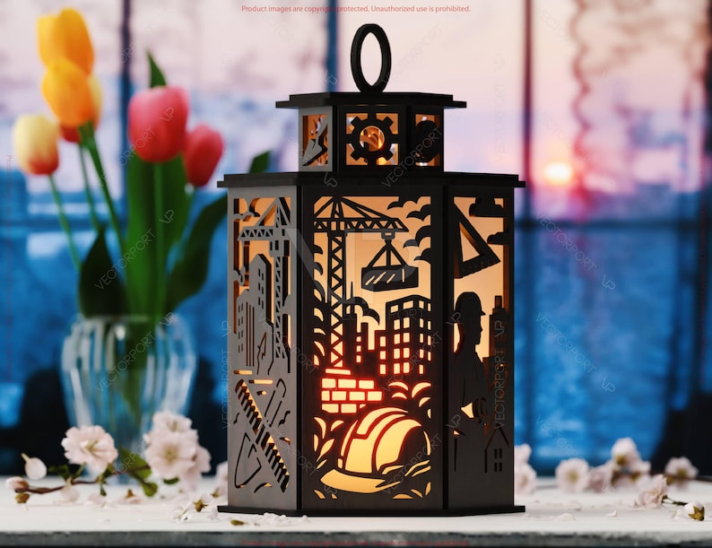 May include: Black laser-cut lantern with construction-themed cutouts, including a crane, buildings, and a hard hat. The lantern has a handle and is illuminated from within, creating a warm glow. The background features flowers and a window.