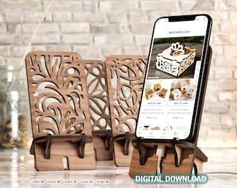 May include: Four wooden phone stands with intricate floral designs. The stands are made of light brown wood and have a cutout design. The stands are on a table with a brick wall in the background. A smartphone is in one of the stands, showing a website with a product listing for a ring box. The text on the website says "Mother's Day".