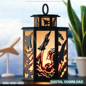 May include: Black laser-cut lantern with an aviation theme. The lantern features cut-out designs of aeroplanes and rockets, with a warm light emanating from within. The lantern has a handle at the top and the words "DIGITAL DOWNLOAD" at the bottom.