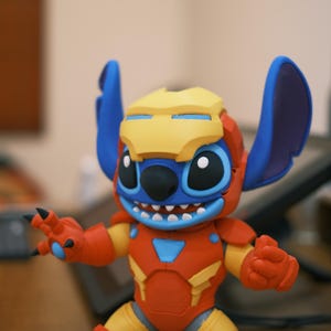 May include: A colorful figurine of Stitch in an Iron Man-inspired suit. The figure features a yellow helmet, blue ears, and a red and yellow suit. The figure has a smiling expression and is posed with its arms outstretched.