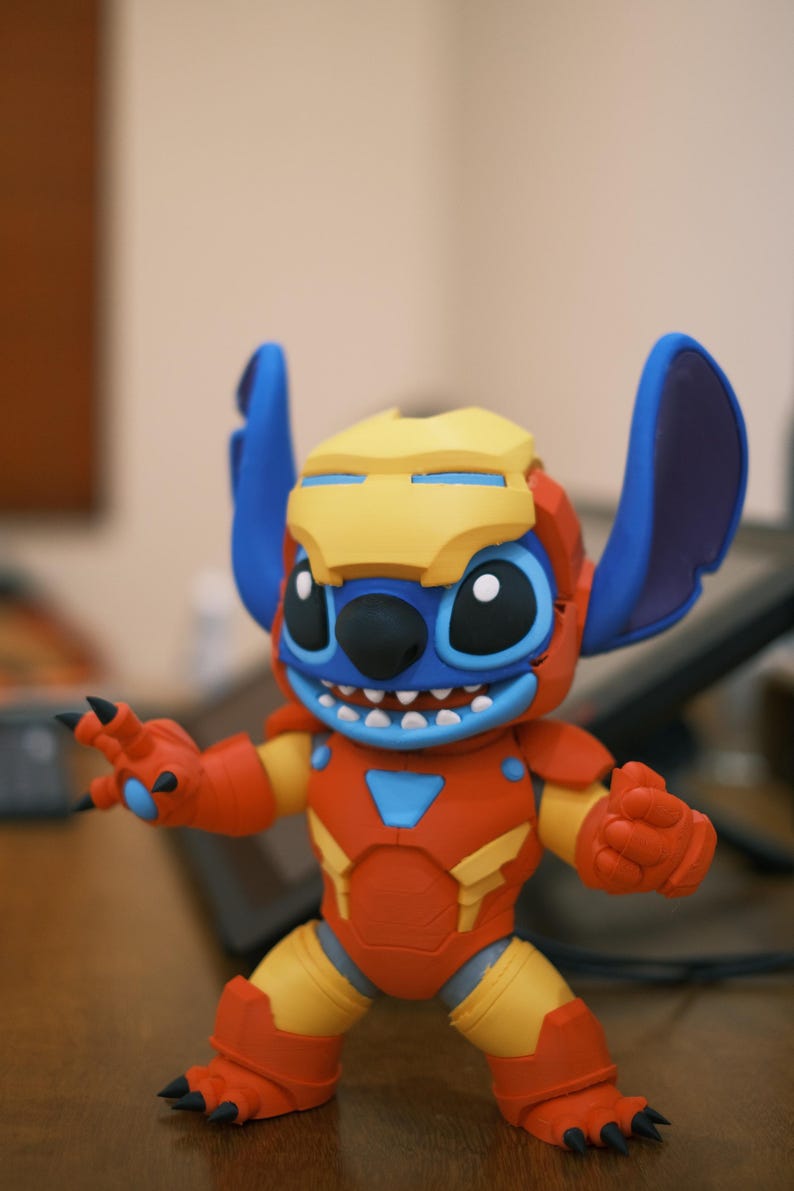 May include: A colorful figurine of Stitch in an Iron Man-inspired suit. The figure features a yellow helmet, blue ears, and a red and yellow suit. The figure has a smiling expression and is posed with its arms outstretched.