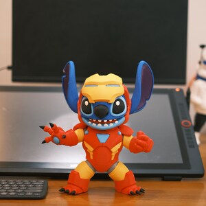 May include: A figurine of Stitch, the Disney character, in an Iron Man suit. The figure is primarily red and yellow, with blue ears and head. The figure is posed with its arms out, and has a smiling expression.