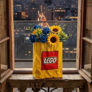 May include: A bright yellow paper bag featuring a red square with the white and black LEGO logo, filled with a bouquet of artificial blue roses, yellow sunflowers, and green succulent plants, positioned against a blurred city skyline background.