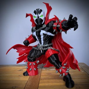 May include: A Spawn action figure featuring a black suit with white chest markings, a flowing red cape, and silver spiked gauntlets and leg guards. The character wears a silver chain belt with a skull buckle and has a white mask with glowing green eyes.