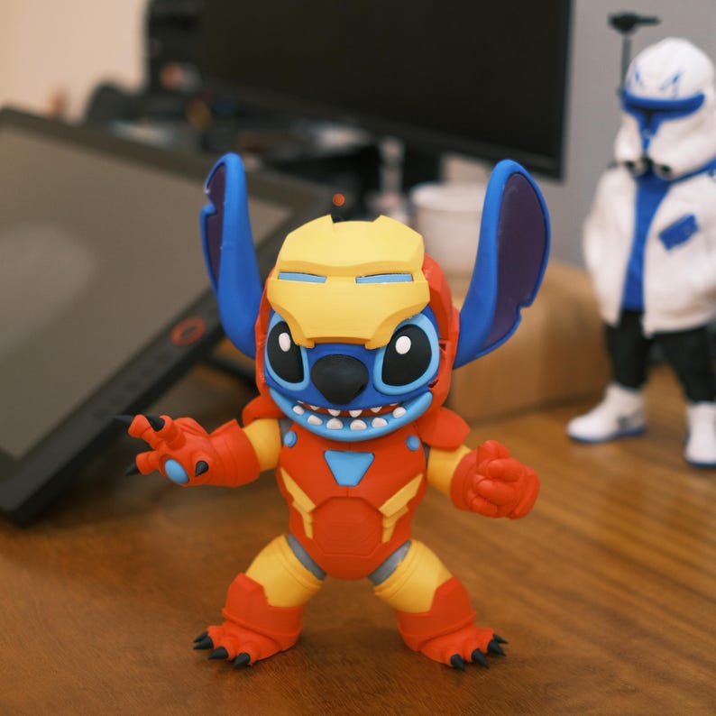 May include: A figurine of Stitch, the Disney character, in an Iron Man suit. The figure is primarily red, yellow, and blue, with Stitch's signature large ears and a helmet. The figure is posed with one arm extended. Another figurine is in the background.
