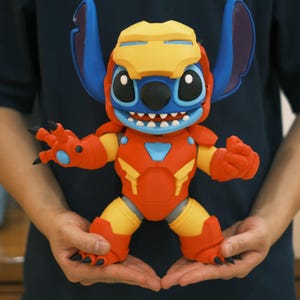 May include: A colorful figurine of Stitch, the Disney character, in an Iron Man suit. The figure is primarily red, yellow, and blue, with detailed armor and Stitch's signature large ears. The figure is held in open hands.