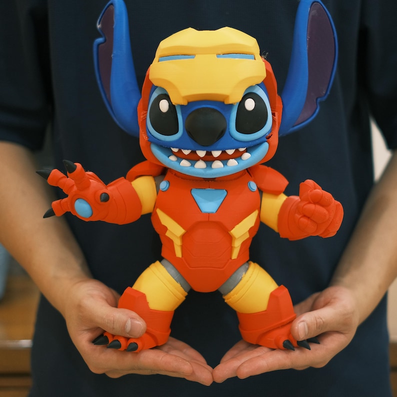 May include: A colorful figurine of Stitch, the Disney character, in an Iron Man suit. The figure is primarily red, yellow, and blue, with detailed armor and Stitch's signature large ears. The figure is held in open hands.