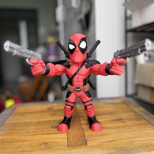 May include: A red and black Deadpool action figure holding two silver toy guns. The figure has a black mask with white eyes and a brown belt with a logo. Two black swords are on the figure's back.