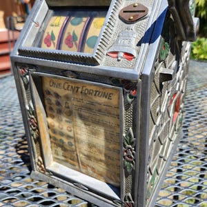 May include: A vintage metal One Cent Fortune trade stimulator machine featuring a coin slot, a side lever, and three spinning reels with fruit symbols. The front panel displays printed text and a bell icon with the year 1776 embossed in silver.
