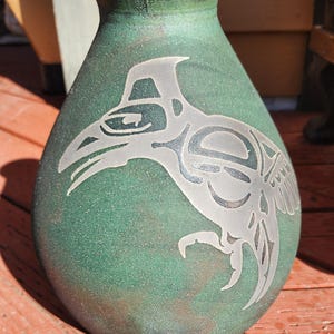 May include: A green ceramic vase with a silver-colored design of a bird with outstretched wings. The vase has a round body and a narrow neck.