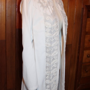 May include: A white long-sleeved jacket with a decorative lace trim down the front. The lace is a delicate white and has a floral pattern.
