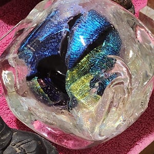 May include: A clear glass bowl with iridescent blue and green accents. The bowl has a unique, organic shape and contains colorful, textured glass pieces. The glass reflects light, creating a shimmering effect. The bowl is a decorative item.