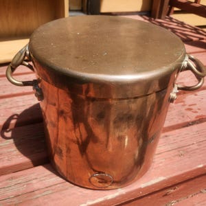 May include: A cylindrical copper pot with a matching lid and two side handles. The metal surface shows a warm, metallic finish with signs of natural oxidation and wear. The pot features a flat top lid and two curved handles attached to the sides.