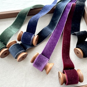 Op de afbeelding: Zes spoelen met fluwelen lint in verschillende kleuren, waaronder groen, blauw, paars en bordeauxrood. De spoelen zijn van hout en hebben een natuurlijke afwerking.