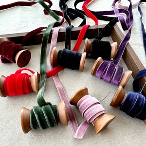 May include: Assortment of spools of velvet ribbon in various colors, including red, green, purple, pink, and blue. The spools are made of wood and have a natural finish. The ribbon is wound around the spools and is ready to be used for crafting or decorating.